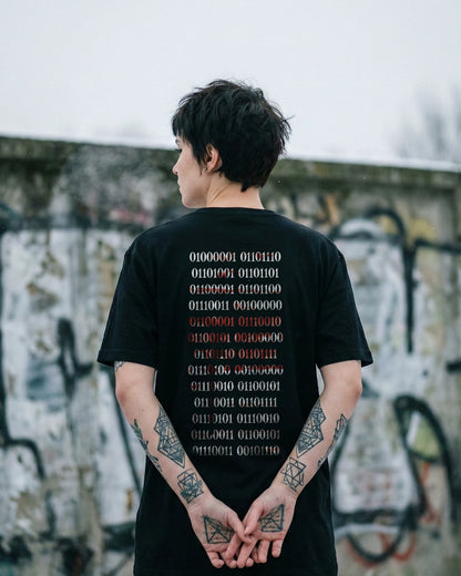 Urbanes Lifestyle-Bild eines schwarzen Natura Umbra Streetwear T-Shirts. Die Person steht mit dem Rücken zur Kamera vor einer Graffiti-Wand und präsentiert das dystopische Binärcode-Design.
