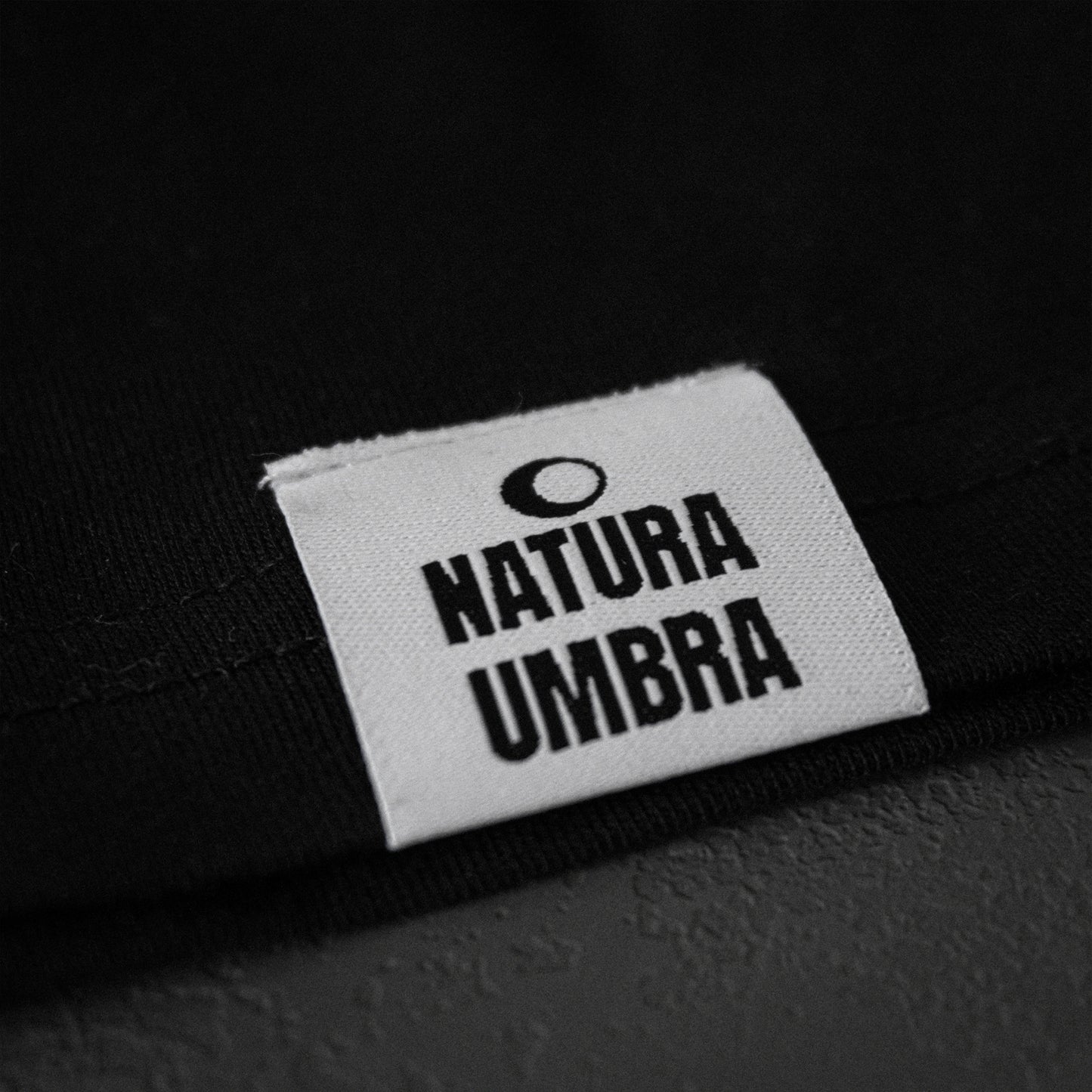 Schwarzes Unisex T-Shirt von Natura Umbra – Detailansicht des gewebten Markenetiketts am linken Saum.