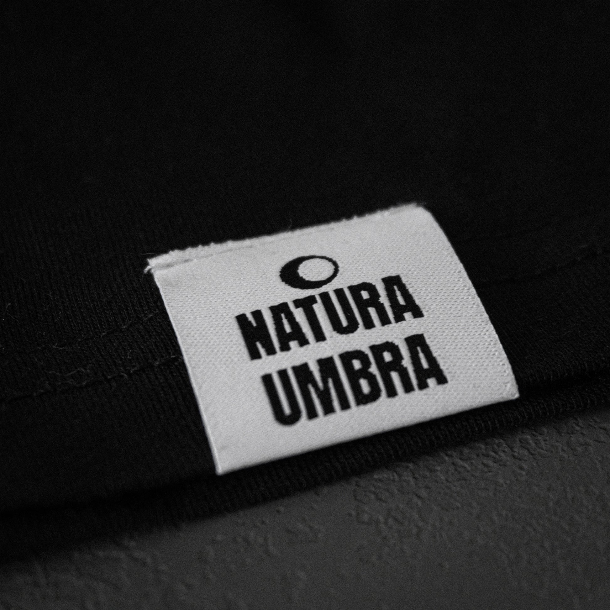 Schwarzes Unisex T-Shirt von Natura Umbra – Detailansicht des gewebten Markenetiketts am linken Saum.