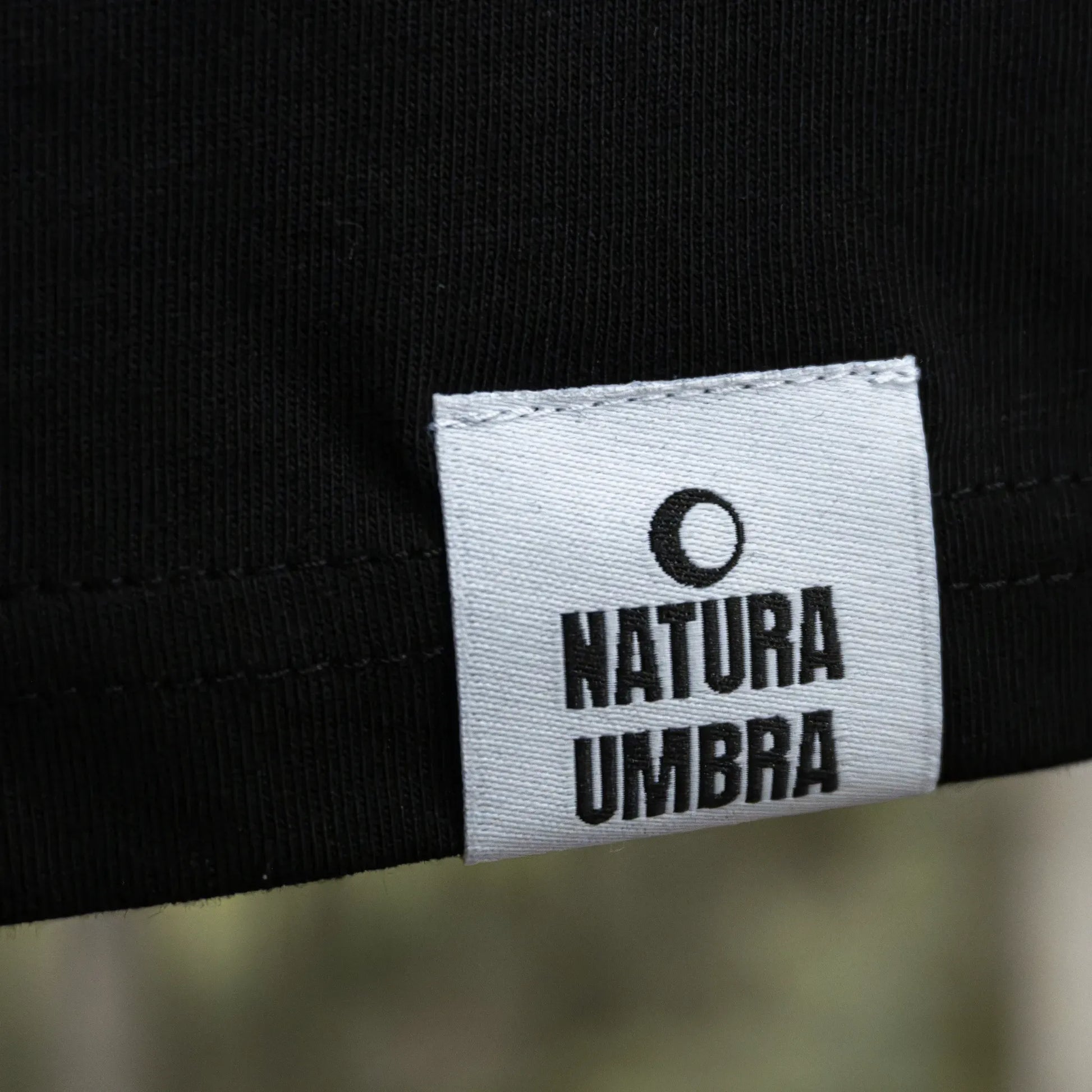 Schwarzes Unisex T-Shirt von Natura Umbra – Detailansicht des gewebten Markenetiketts am linken Saum.