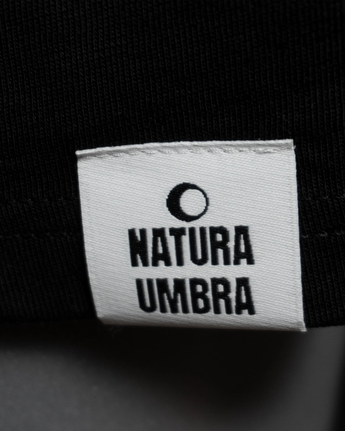 Detailaufnahme des Webetiketts am Saum des T-Shirts in Schwarz – dezentes Branding-Element von Natura Umbra.