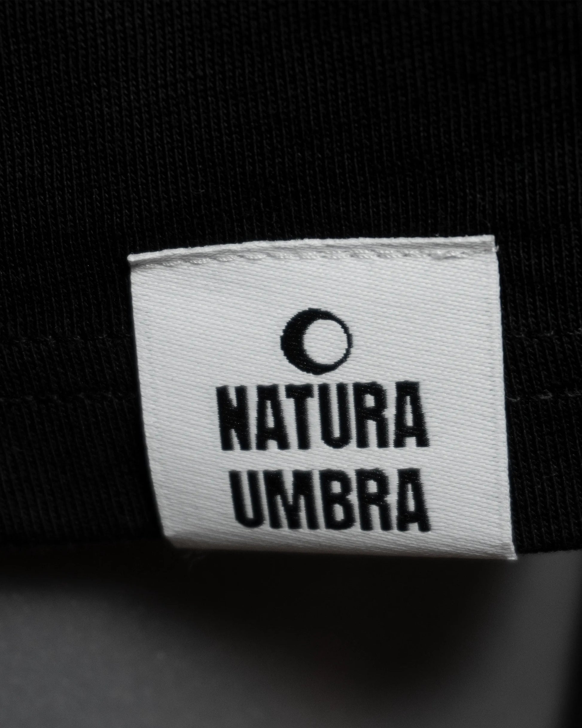 Detailaufnahme des Webetiketts am Saum des T-Shirts in Schwarz – dezentes Branding-Element von Natura Umbra.