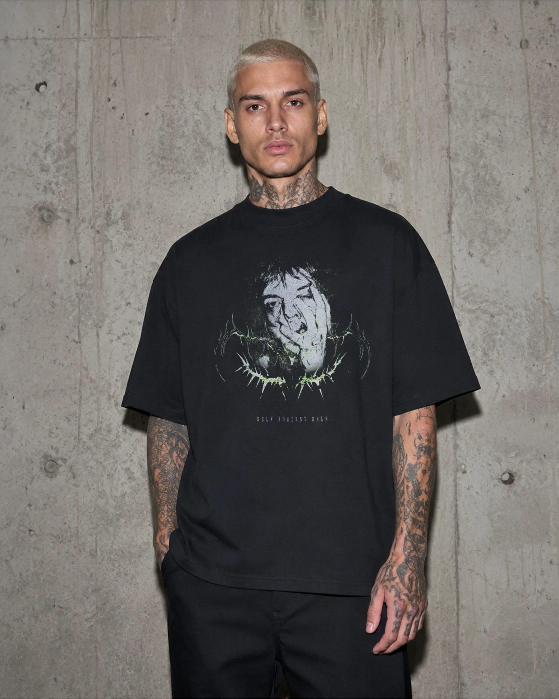 Model trägt schwarzes SELF AGAINST SELF Oversized T-Shirt vor einer Betonwand.